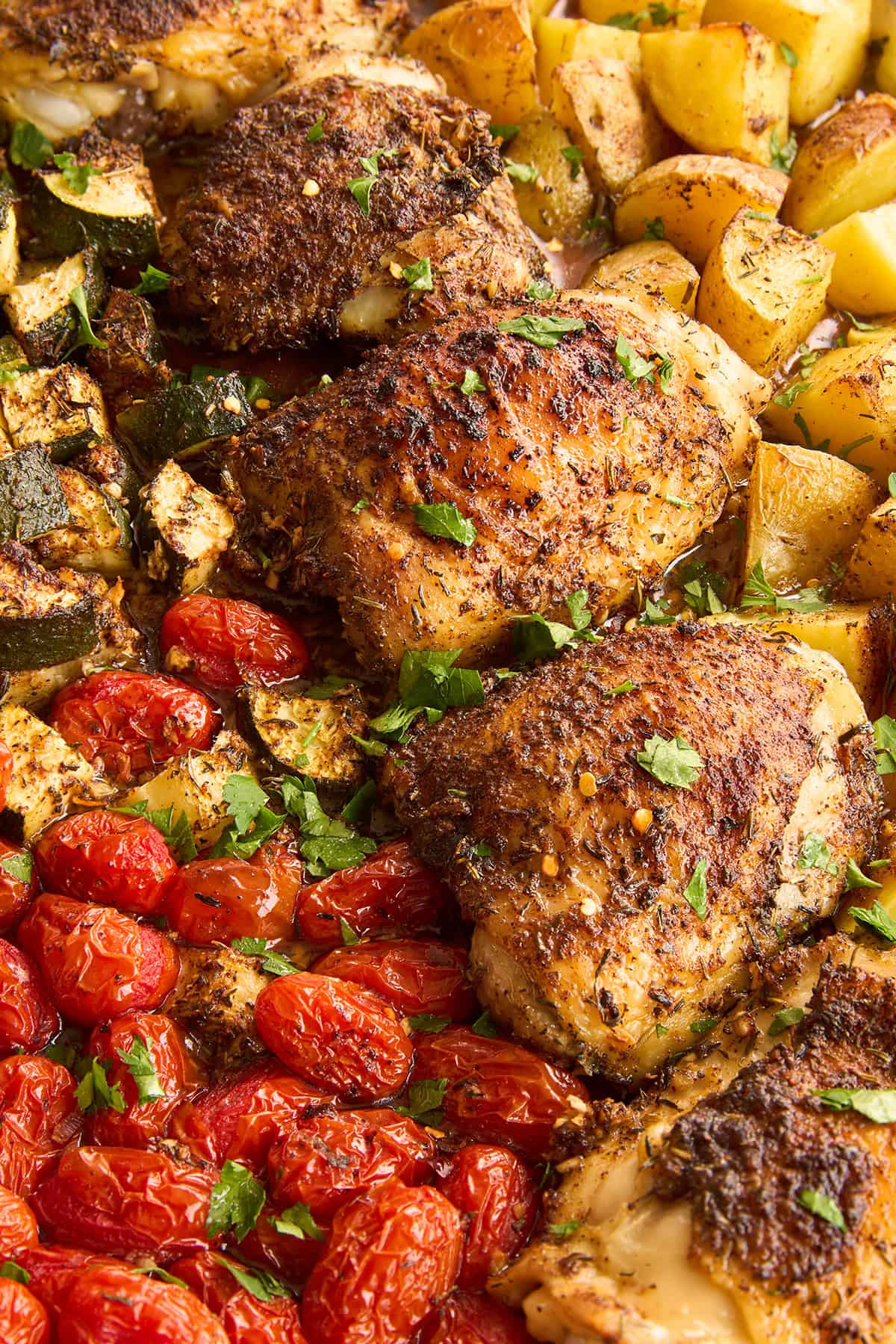 Seasoned roasted chicken thighs on a sheet pan with golden roasted potatoes, cherry tomatoes, and zucchini, garnished with chopped fresh herbs.