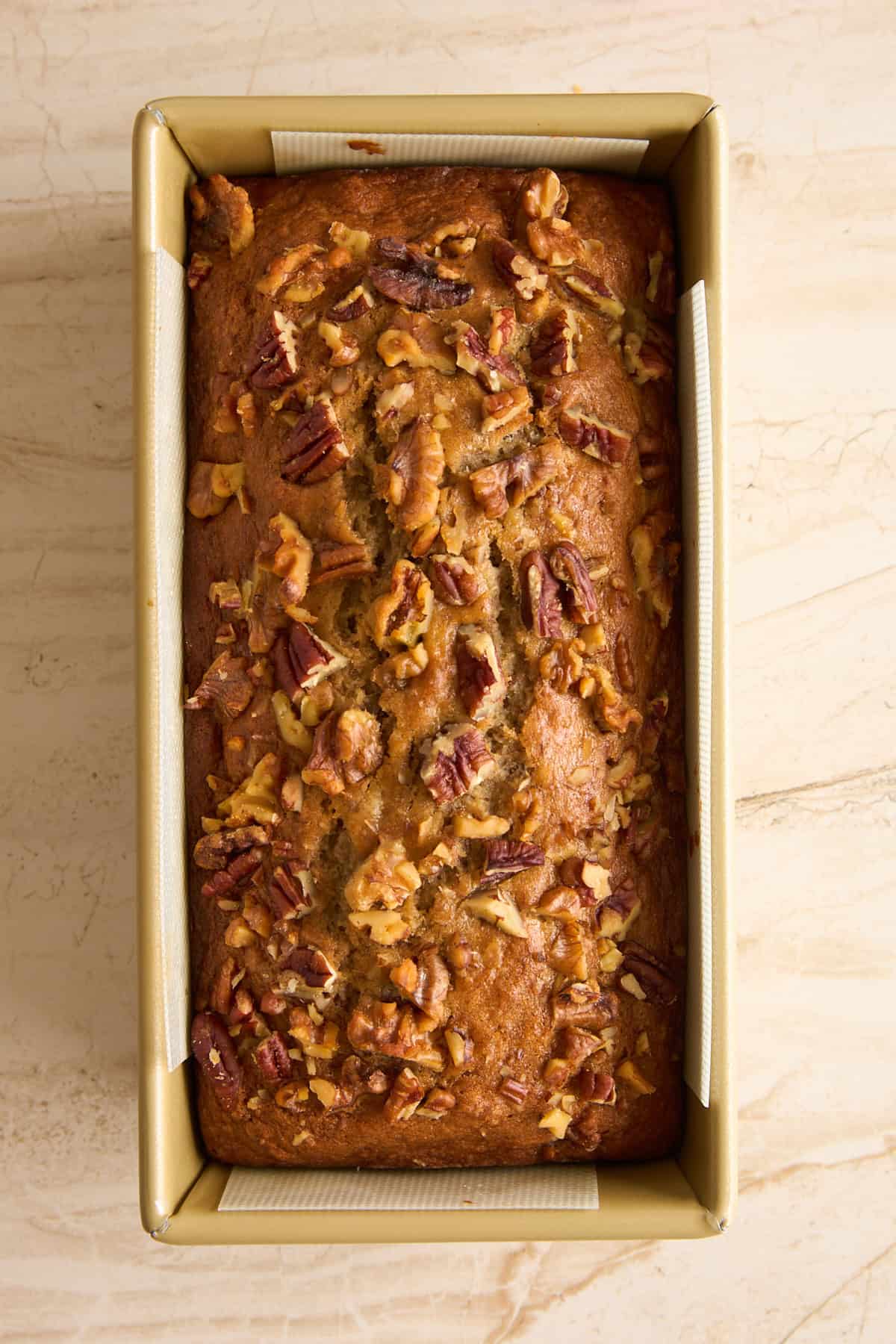 A loaf of banana bread topped with chopped walnuts sits in a rectangular, gold-colored baking pan on a light marble surface.