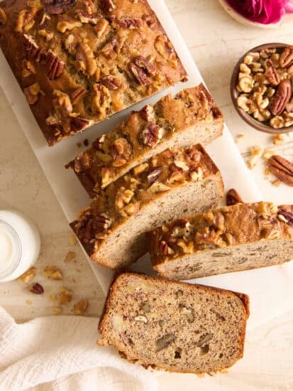A loaf of banana nut bread, topped with chopped walnuts and pecans, sits on a white cutting board. Several slices are cut, and a bowl of mixed nuts is nearby. A glass of milk and a pink flower are also visible.