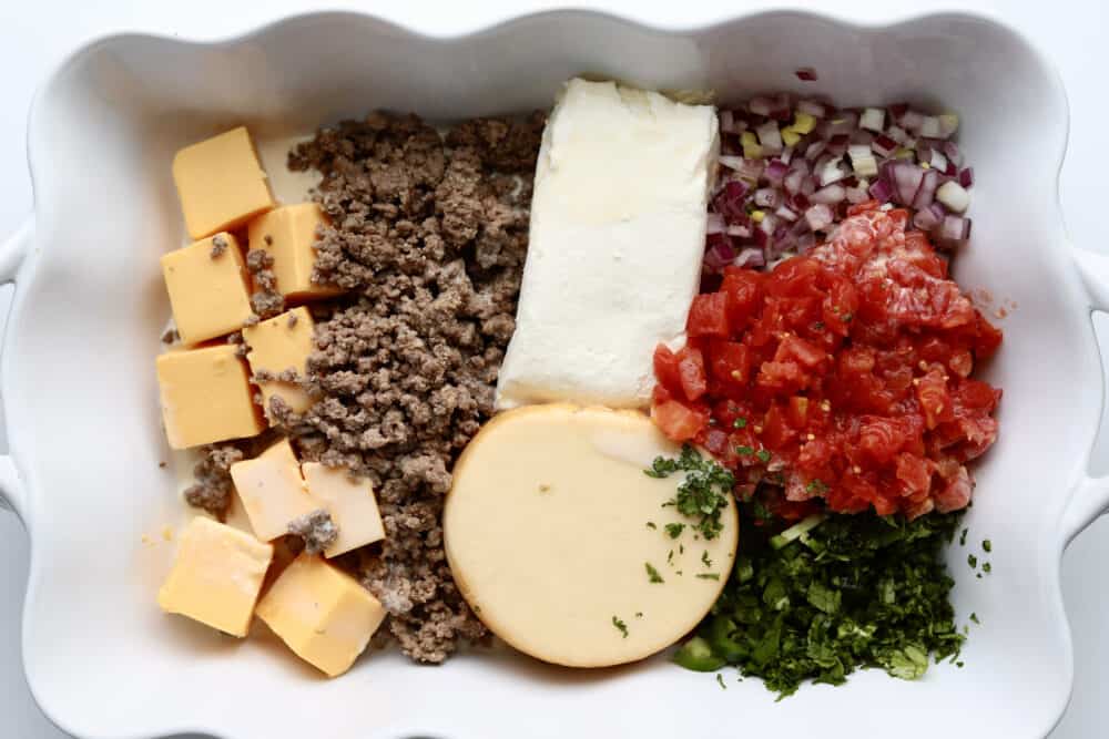 overhead image of cubed velveeta cheese, lean beef, cream cheese, gouda cheese, diced red onion, canned tomatoes, and cilantro in a white baking dish
