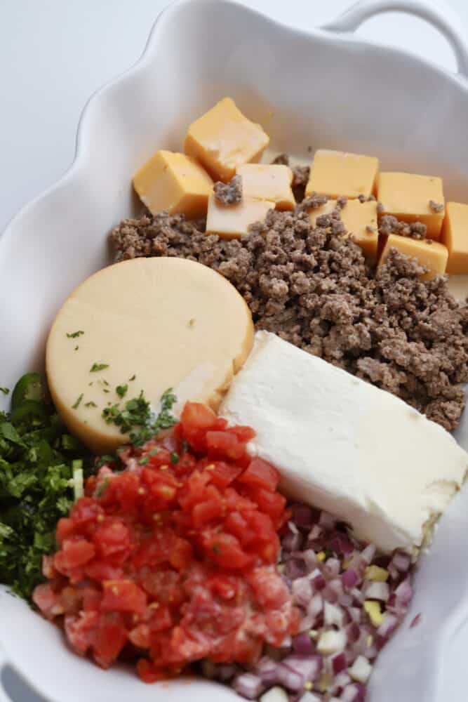 ingredients for homemade queso fundido dip in a white baking dish including chopped cilantro, canned tomatoes, rex onions, gouda cheese, cream cheese, lean beef, and velveeta cheese