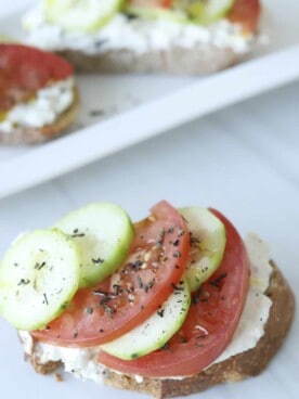 a piece of feta toast topped with sliced tomatoes, cucumbers, and herbs