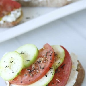 a piece of feta toast topped with sliced tomatoes, cucumbers, and herbs
