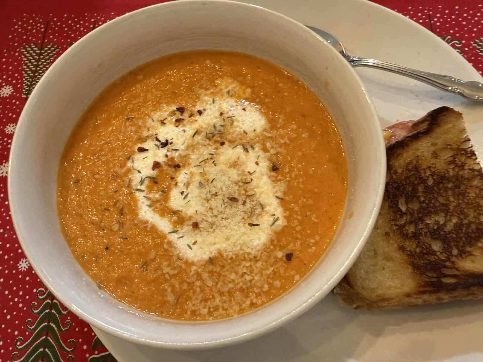 A bowl of tomato soup topped with cream and herbs sits on a white plate next to a grilled cheese sandwich; the tablecloth has a festive red and green pattern.