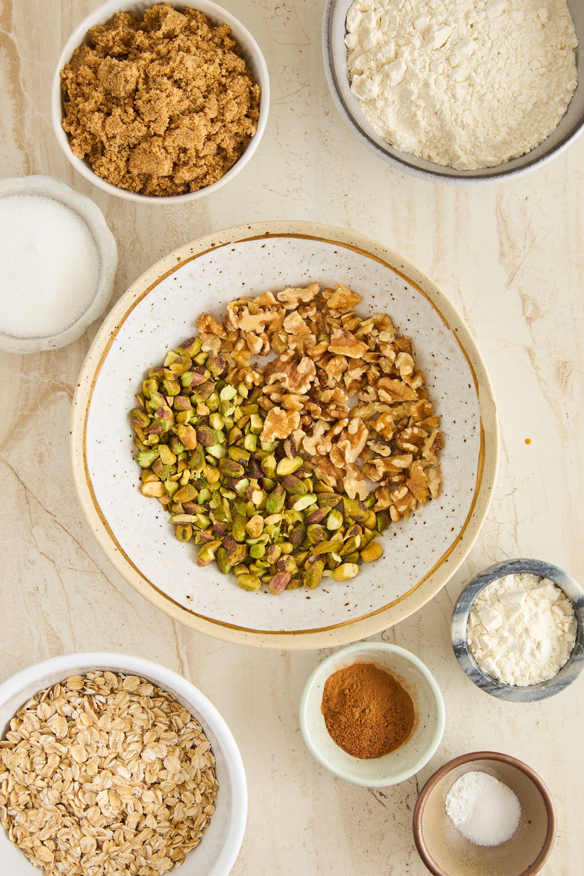 A variety of baking ingredients in bowls on a light surface, including walnuts, pistachios, brown sugar, flour, oats, cinnamon, cornstarch, salt, and granulated sugar.