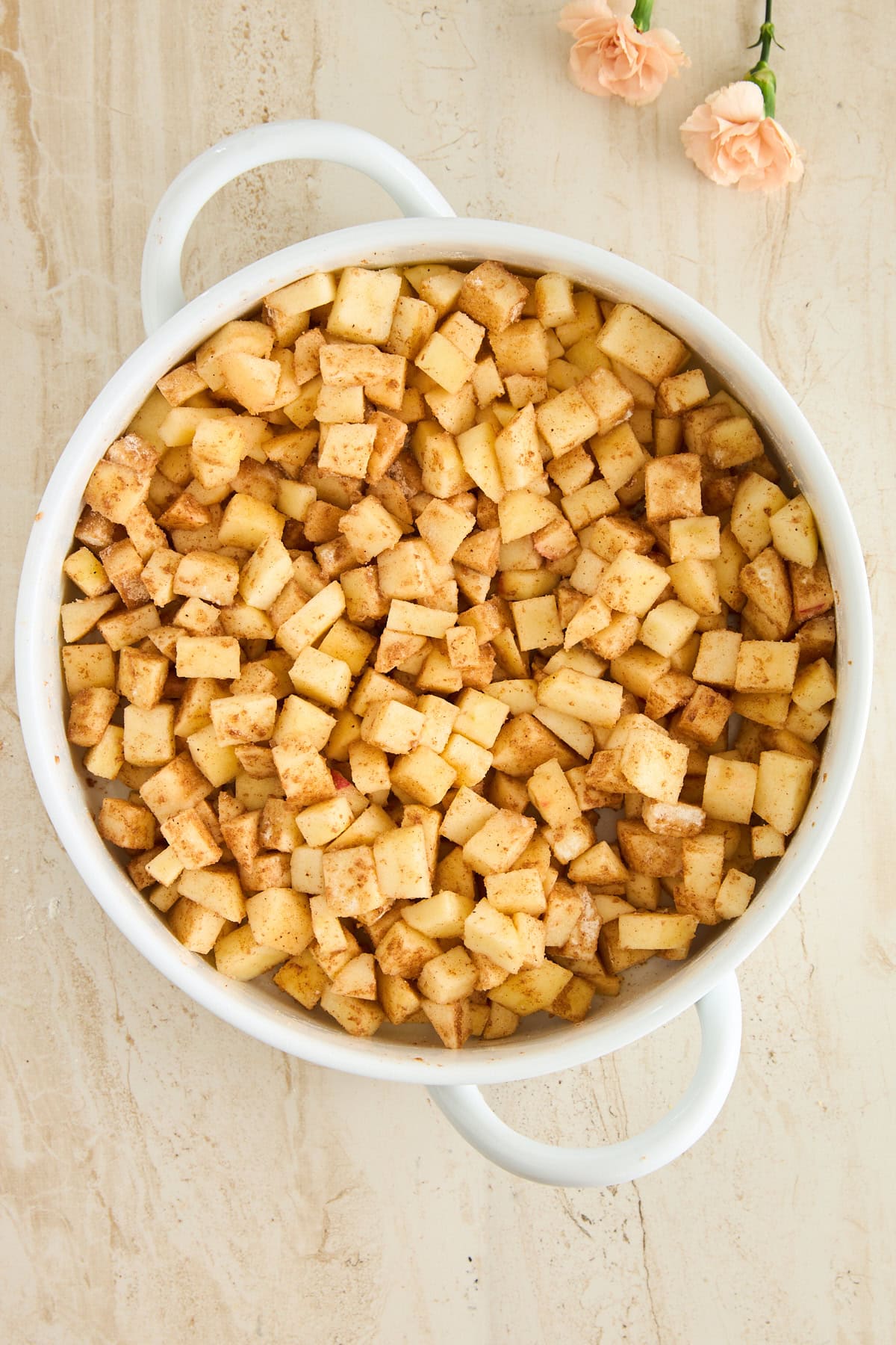 A white pot filled with diced apples coated in cinnamon sits on a light beige surface, with two light pink flowers placed nearby. Ingredients for Baklava Apple Crisp.