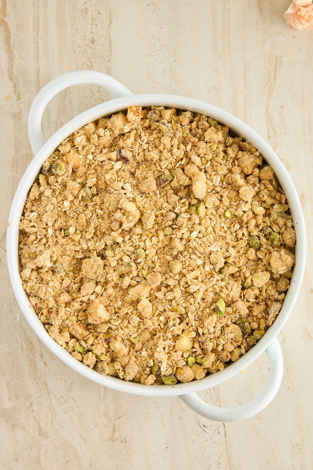 A round white baking dish with handles filled with a golden-brown oat and nut crumble topping, set on a light beige textured surface.
