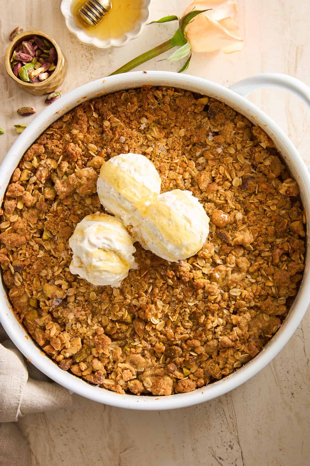 A round white dish filled with golden-brown crumble, topped with three scoops of vanilla ice cream drizzled with honey. A small bowl of pistachios, honey dipper, and a pale rose rest nearby on a light surface.