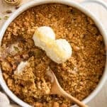 A baked fruit crisp topped with two scoops of vanilla ice cream sits in a round white dish. A wooden spoon is resting in the dish, and some ice cream is melting into the golden, crumbly topping.