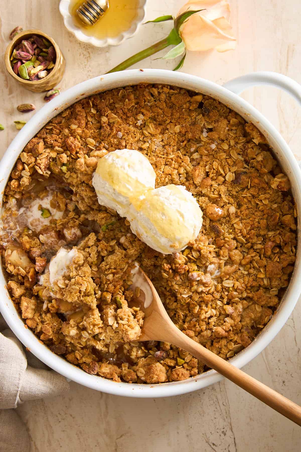 A baked fruit crisp topped with two scoops of vanilla ice cream sits in a round white dish. A wooden spoon is resting in the dish, and some ice cream is melting into the golden, crumbly topping.