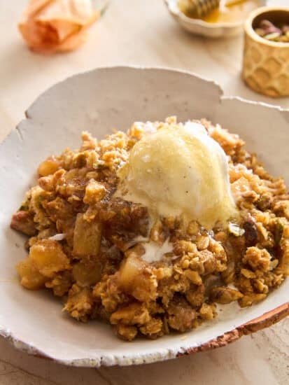 A serving of apple crisp topped with a scoop of melting vanilla ice cream on a rustic ceramic plate, with a honey dipper, small dish, and flower blurred in the background.