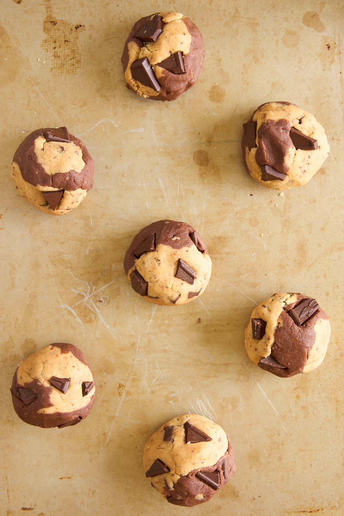 Seven unbaked, swirled chocolate and vanilla cookie dough balls with chocolate chunks are arranged on a lightly scratched baking sheet.