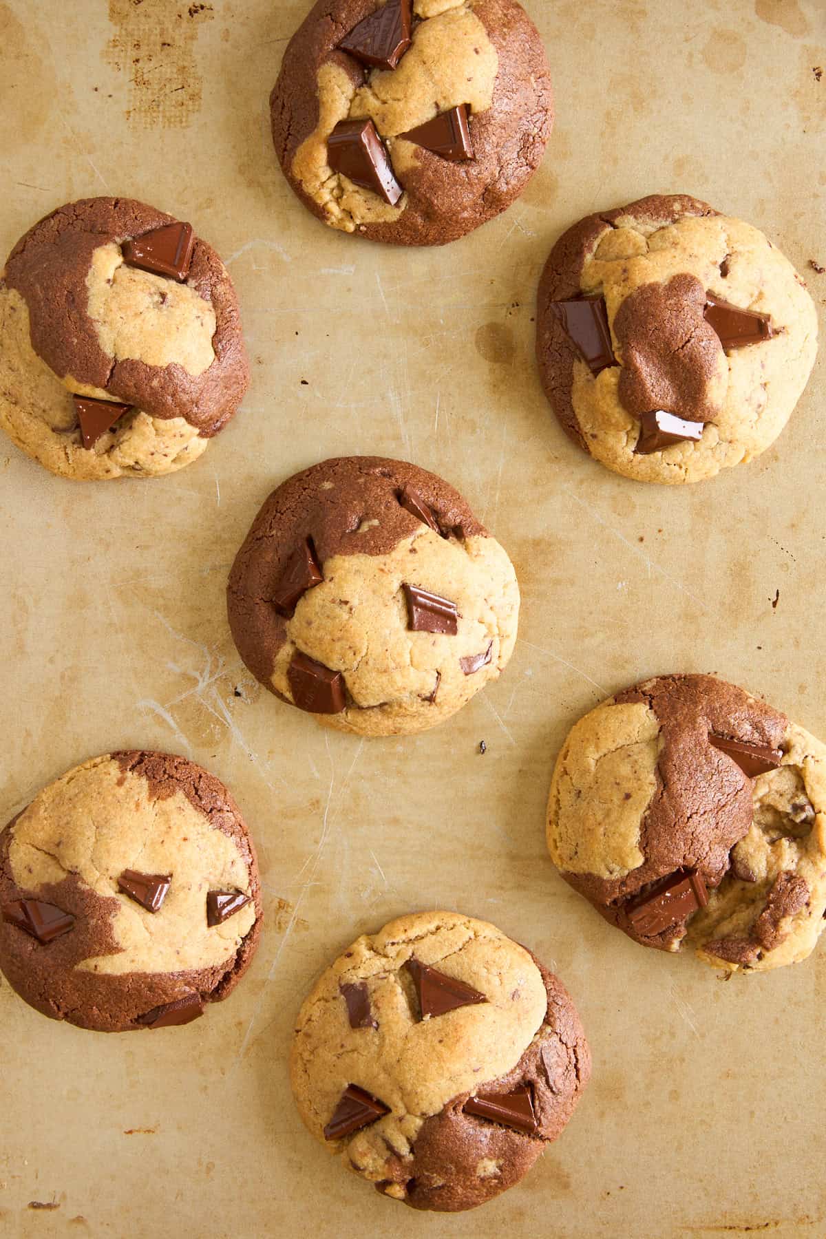 Seven large, marble-patterned cookies with chunks of chocolate are arranged on a tan baking sheet. The cookies have swirls of light and dark dough, and the surface is slightly cracked.
