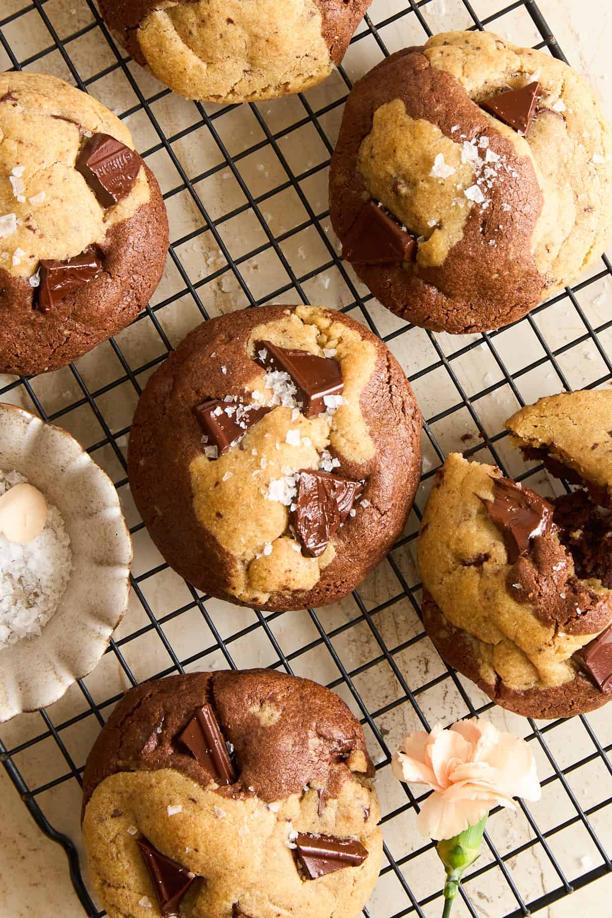 Chocolate chip cookies with sea salt flakes rest on a cooling rack. Some cookies have visible chocolate chunks, and a small bowl of sea salt and a light peach flower sit nearby on the beige surface.