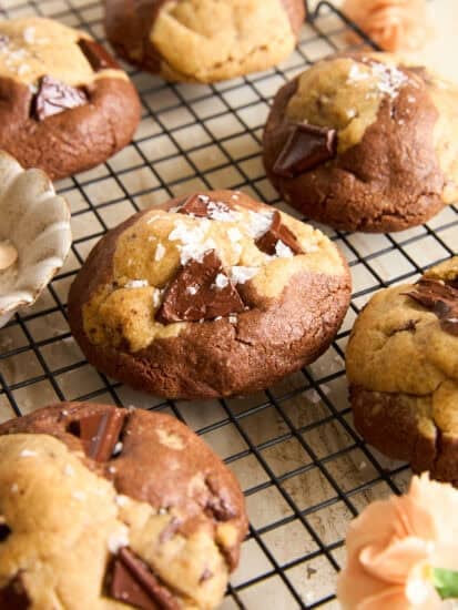 Chocolate chunk cookies with a marbled appearance rest on a black cooling rack. The cookies are sprinkled with flaky sea salt, and there are light orange flowers nearby for decoration.