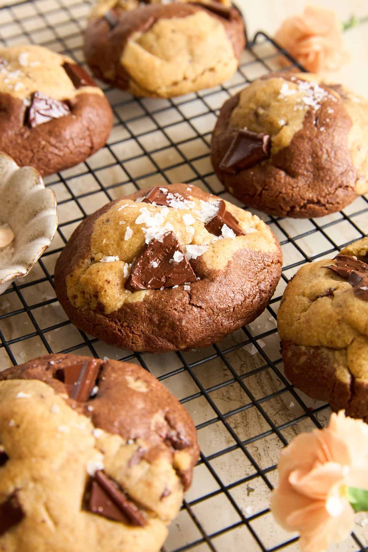 Chocolate chunk cookies with a marbled appearance rest on a black cooling rack. The cookies are sprinkled with flaky sea salt, and there are light orange flowers nearby for decoration.