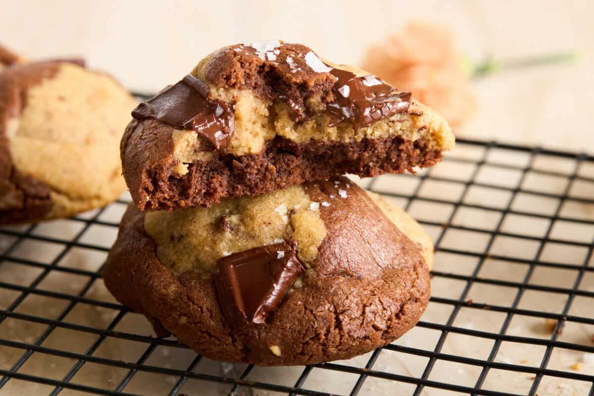 Two chocolate chip cookies, one broken in half to show a soft, gooey center with melted chocolate chunks, stacked on a black wire cooling rack. A blurred flower is visible in the background.