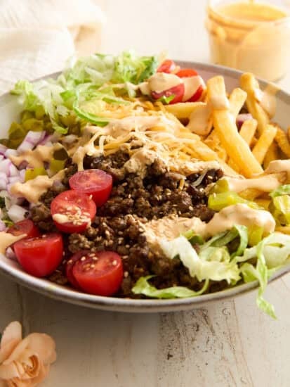 A bowl filled with ground beef, shredded cheese, lettuce, cherry tomatoes, diced onions, pickles, French fries, and drizzled sauce, arranged in sections on a light surface with a small dish of sauce in the background.