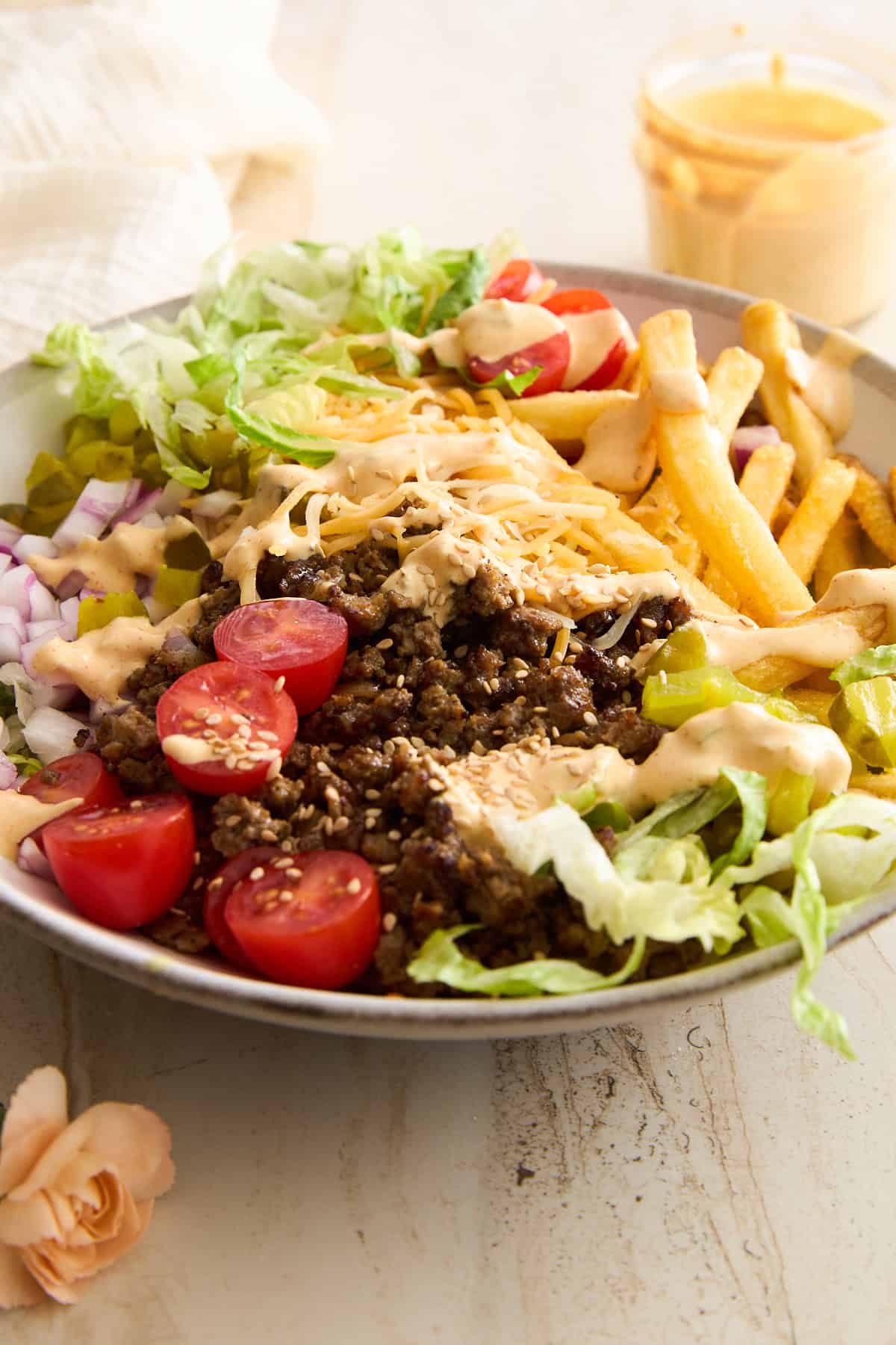A bowl filled with ground beef, shredded cheese, lettuce, cherry tomatoes, diced onions, pickles, French fries, and drizzled sauce, arranged in sections on a light surface with a small dish of sauce in the background.
