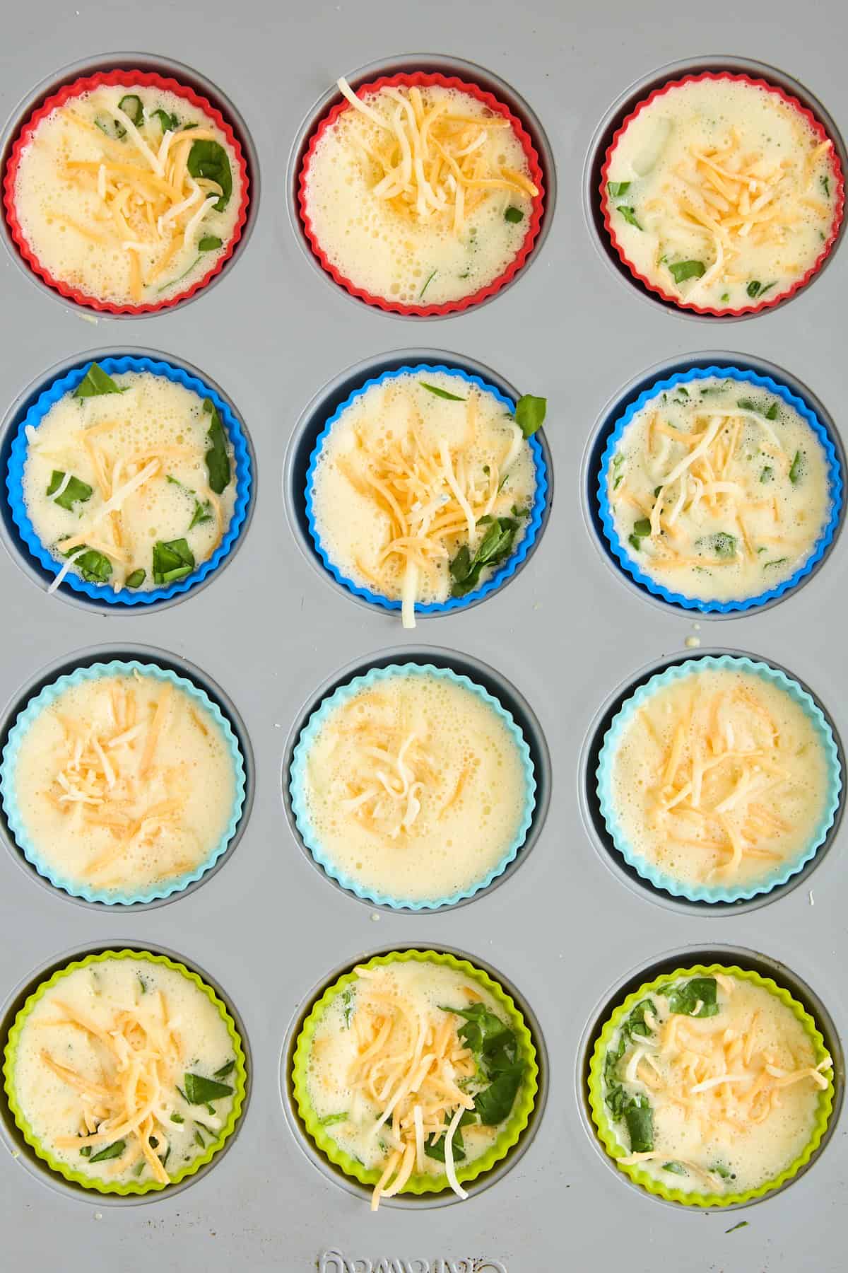 A muffin tin filled with colorful silicone liners holds unbaked egg muffin mixtures topped with shredded cheese and spinach. The mixtures are ready to be baked.