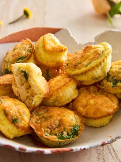 A plate of mini egg muffins with golden-brown tops, filled with spinach and cheese. One muffin is cut open, revealing the fluffy inside and chunky filling. The plate sits on a light surface with flowers in the background.