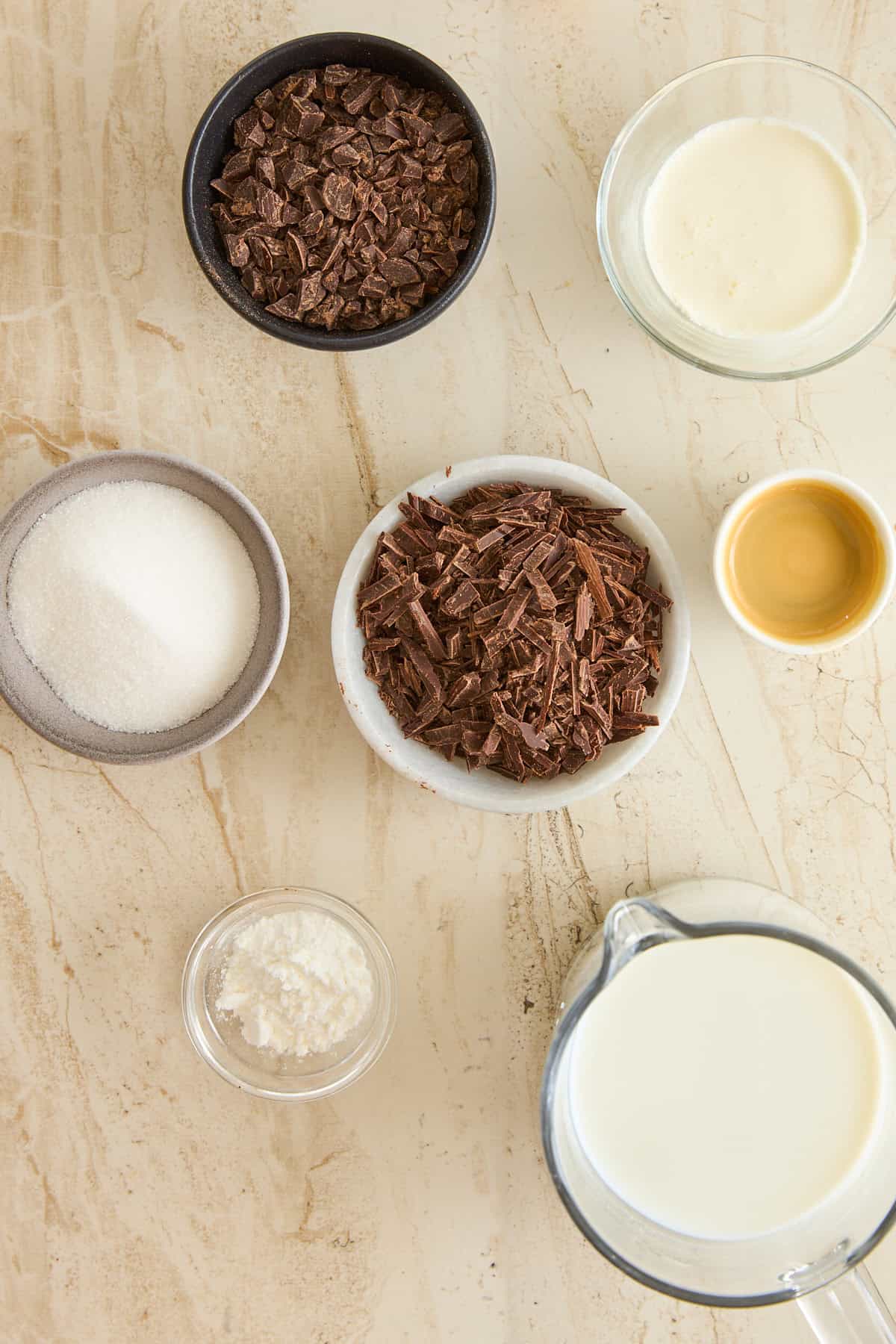 Overhead view of bowls containing chocolate chunks, milk, cream, sugar, a yellow liquid (likely vanilla extract), and a small bowl of cornstarch, arranged on a light-colored marble surface.