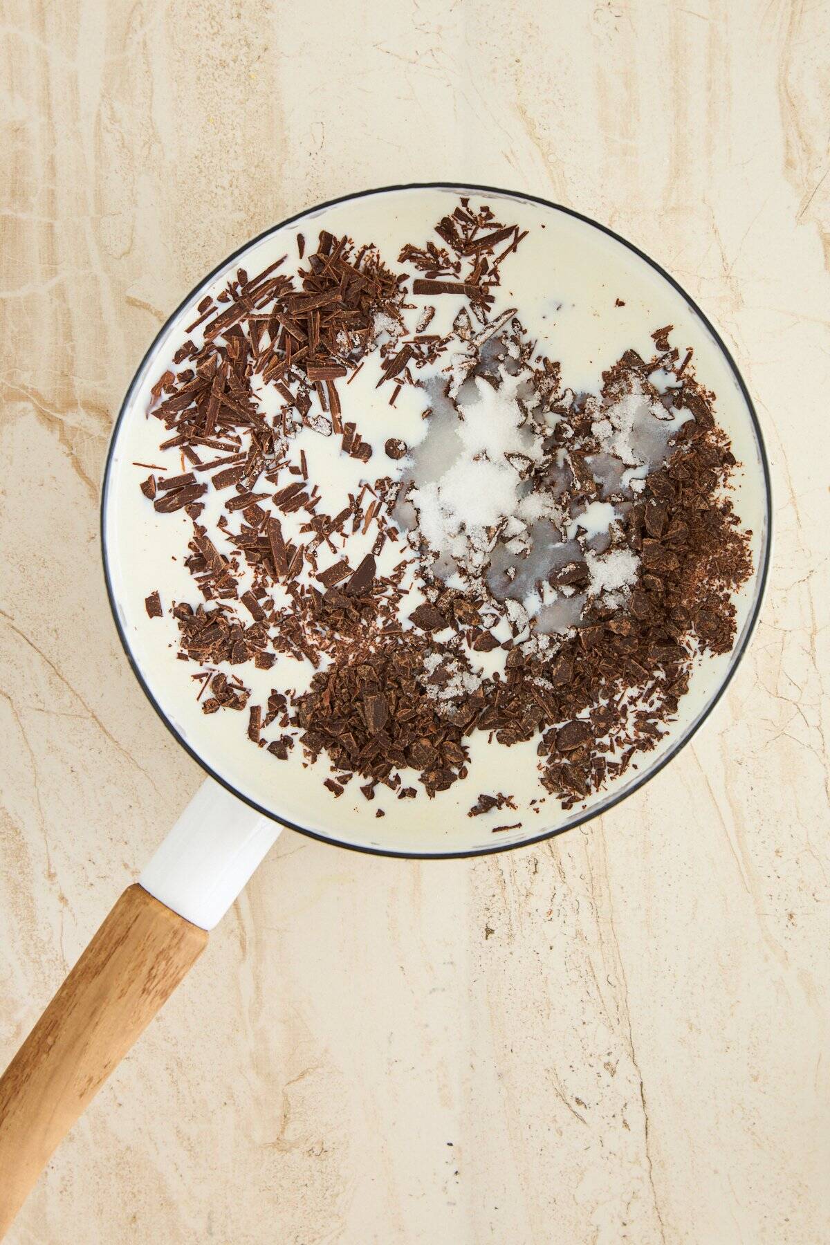 A white saucepan with a wooden handle holds cream, sugar, and chopped dark chocolate, viewed from above on a light beige surface.