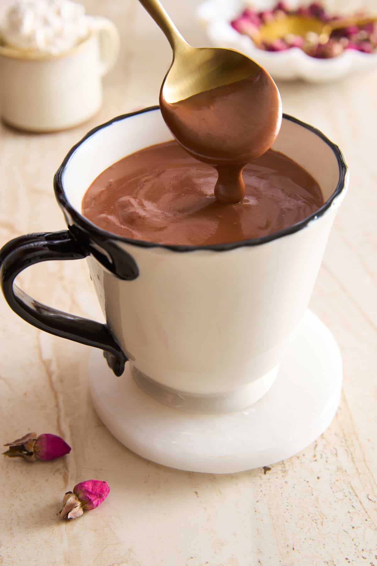 A spoon with thick Italian hot chocolate hovers over a white mug with a black handle, sitting on a white coaster. Rose buds and petals are scattered nearby on a light surface.