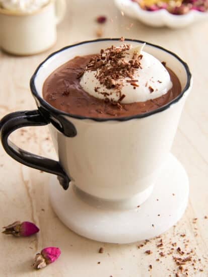 A white mug filled with hot chocolate topped with whipped cream and chocolate shavings, sitting on a marble coaster with dried rosebuds and a second cup blurred in the background.