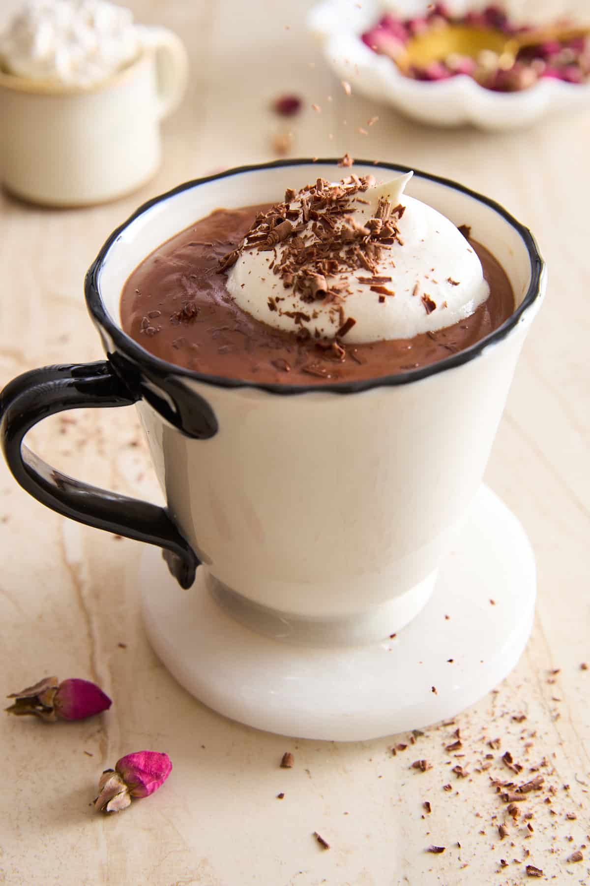 A white mug filled with hot chocolate topped with whipped cream and chocolate shavings, sitting on a marble coaster with dried rosebuds and a second cup blurred in the background.