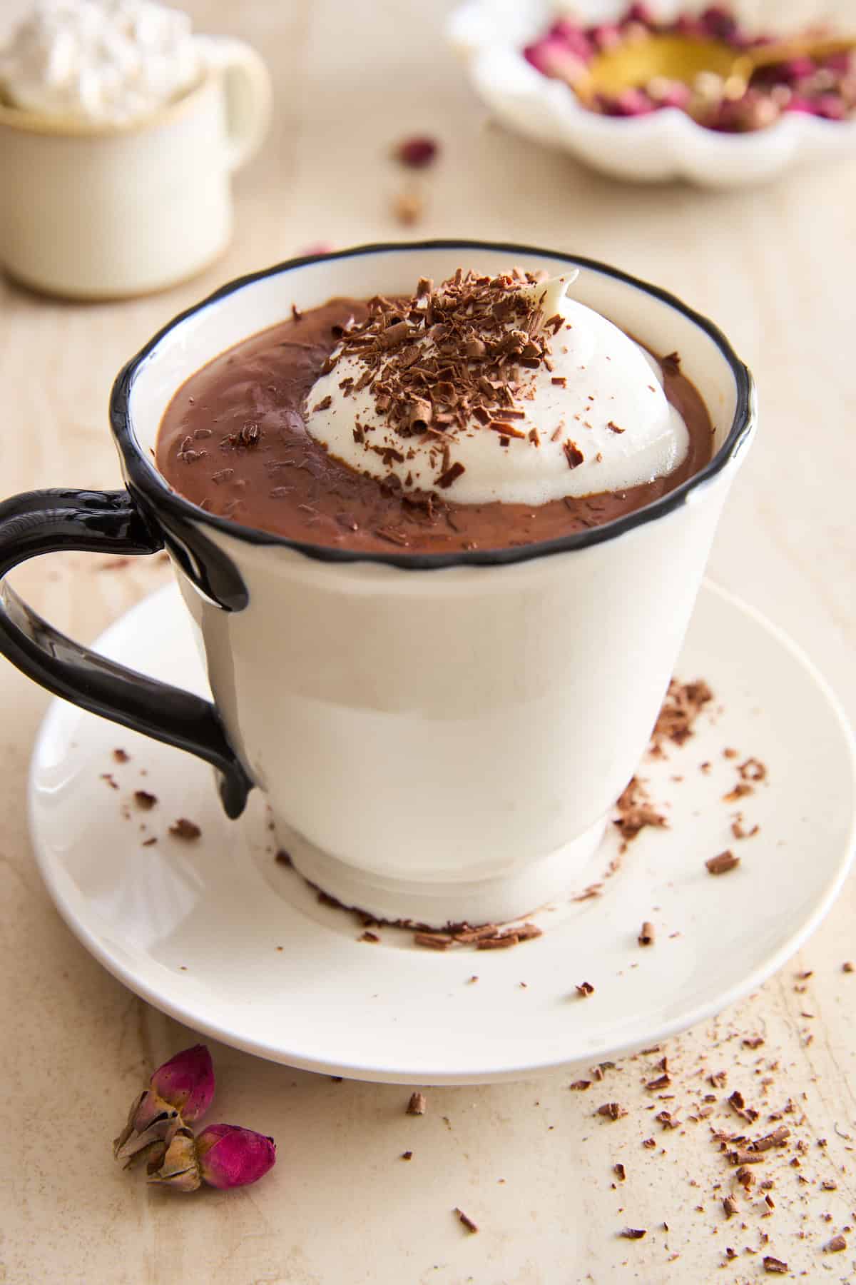 A creamy hot chocolate topped with whipped cream and chocolate shavings in a white mug, sitting on a matching saucer, with rose petals and chocolate shavings scattered nearby.