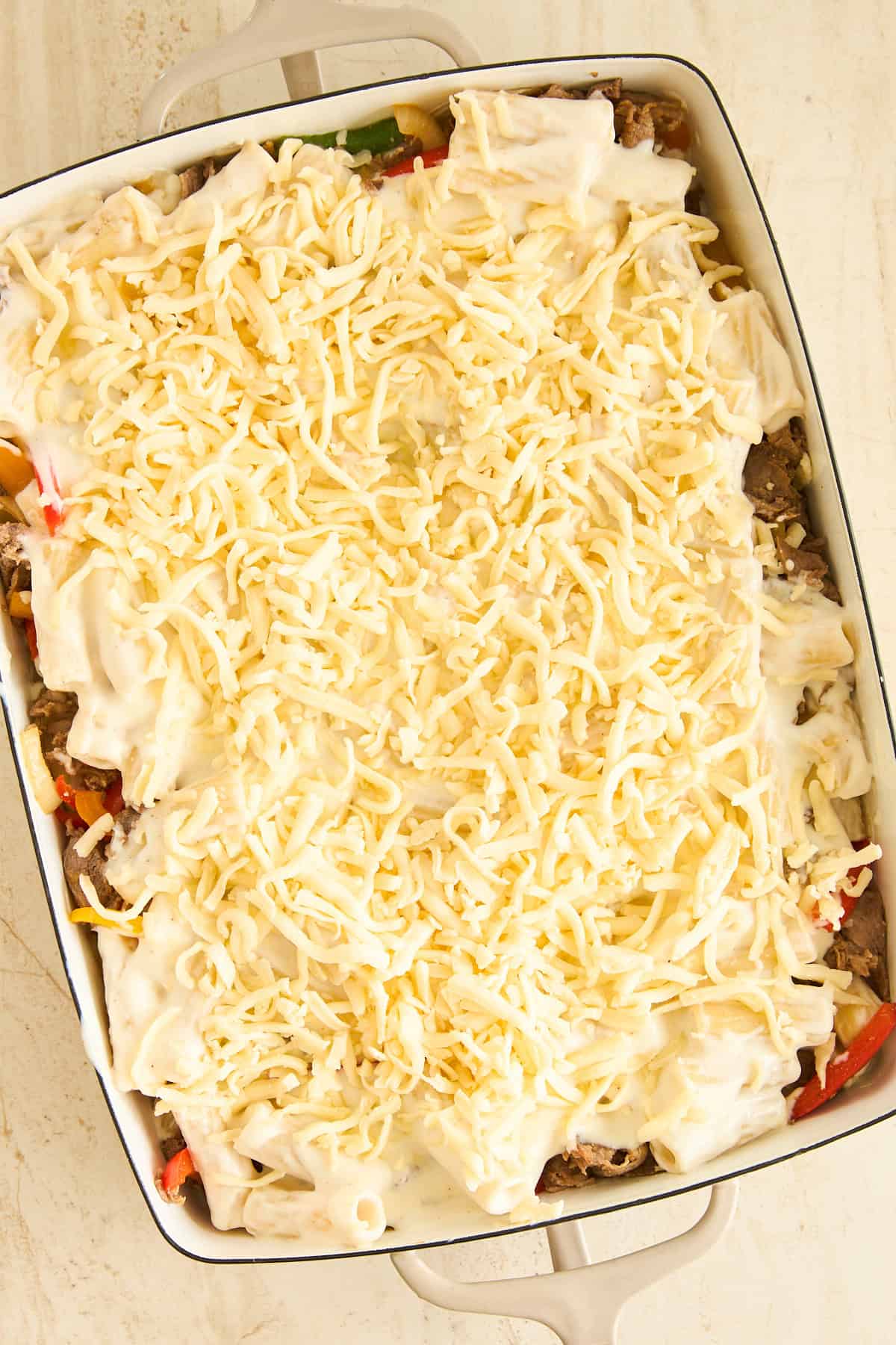 A casserole dish filled with layers of sliced vegetables and meat, topped with a creamy white sauce and a generous amount of shredded cheese, ready to be baked.