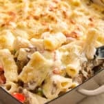 A close-up of a baked pasta casserole in a rectangular dish, featuring rigatoni pasta, melted cheese on top, and visible pieces of vegetables and meat in a creamy sauce. A serving is being scooped out.