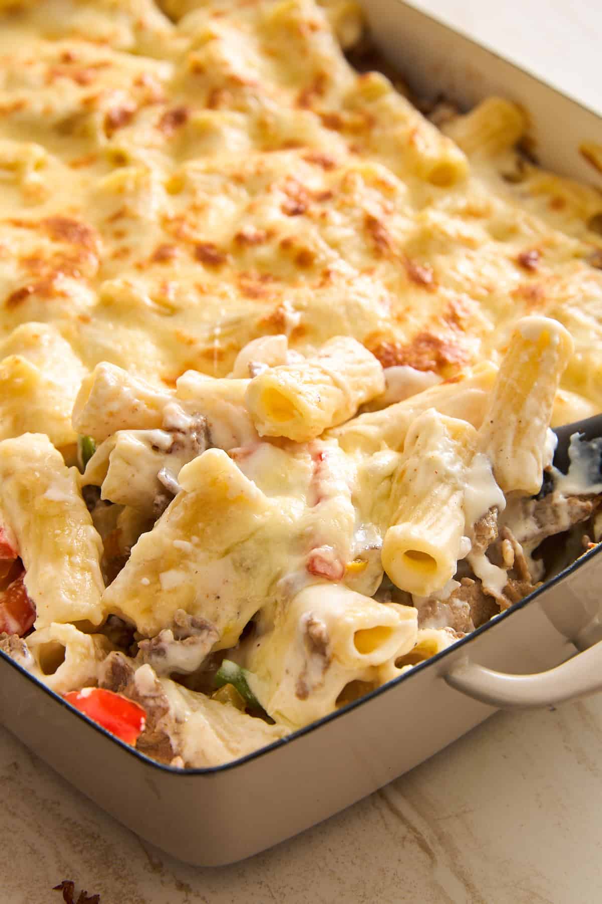 A close-up of a baked pasta casserole in a rectangular dish, featuring rigatoni pasta, melted cheese on top, and visible pieces of vegetables and meat in a creamy sauce. A serving is being scooped out.