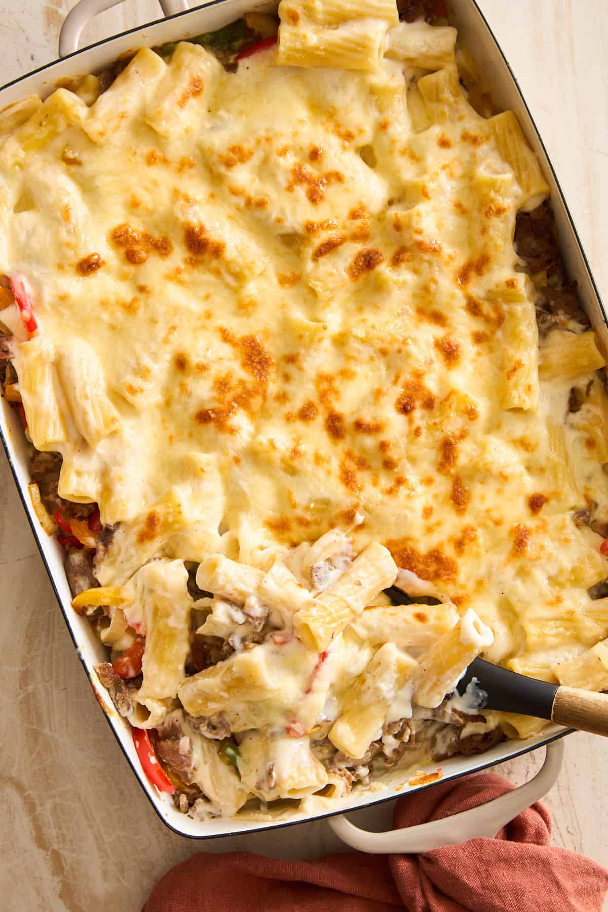 A Philly cheesesteak pasta bake topped with melted, golden-brown cheese in a white rectangular dish. A serving spoon is scooping out a portion, revealing rigatoni and a creamy filling underneath. A red cloth is nearby.
