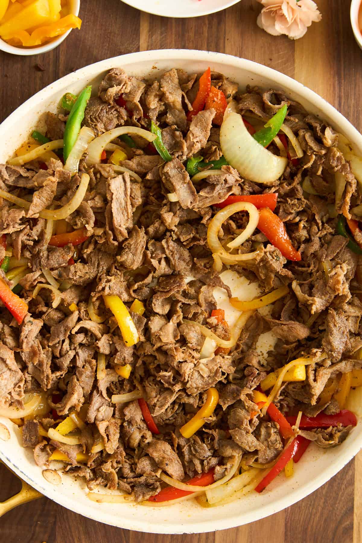 A skillet filled with cooked sliced beef, yellow and red bell peppers, and onions for Philly cheesesteak tacos, on a wooden surface. The colorful vegetables are mixed throughout the meat, creating a vibrant and appetizing dish.