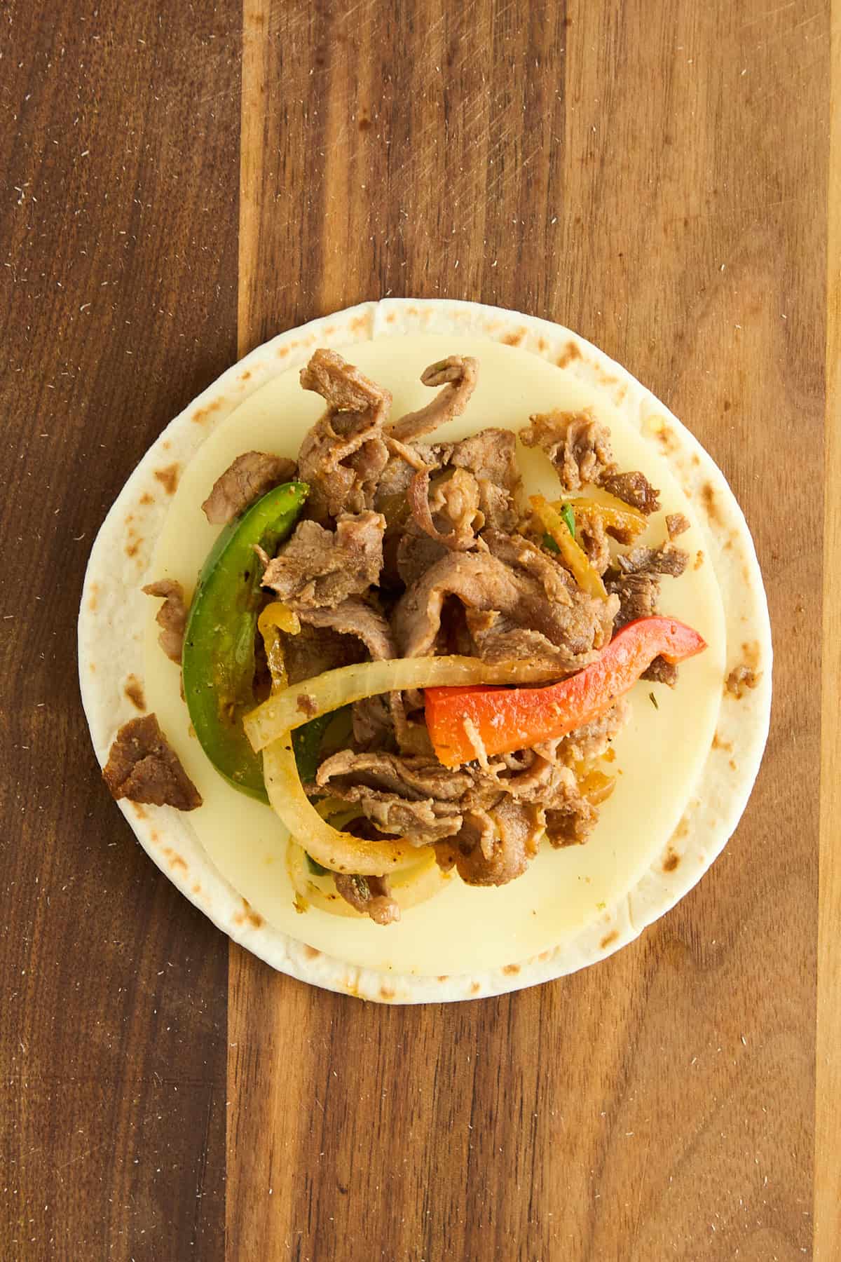 A tortilla topped with melted cheese, sliced cooked beef, green and red bell peppers, and onions sits on a wooden surface.