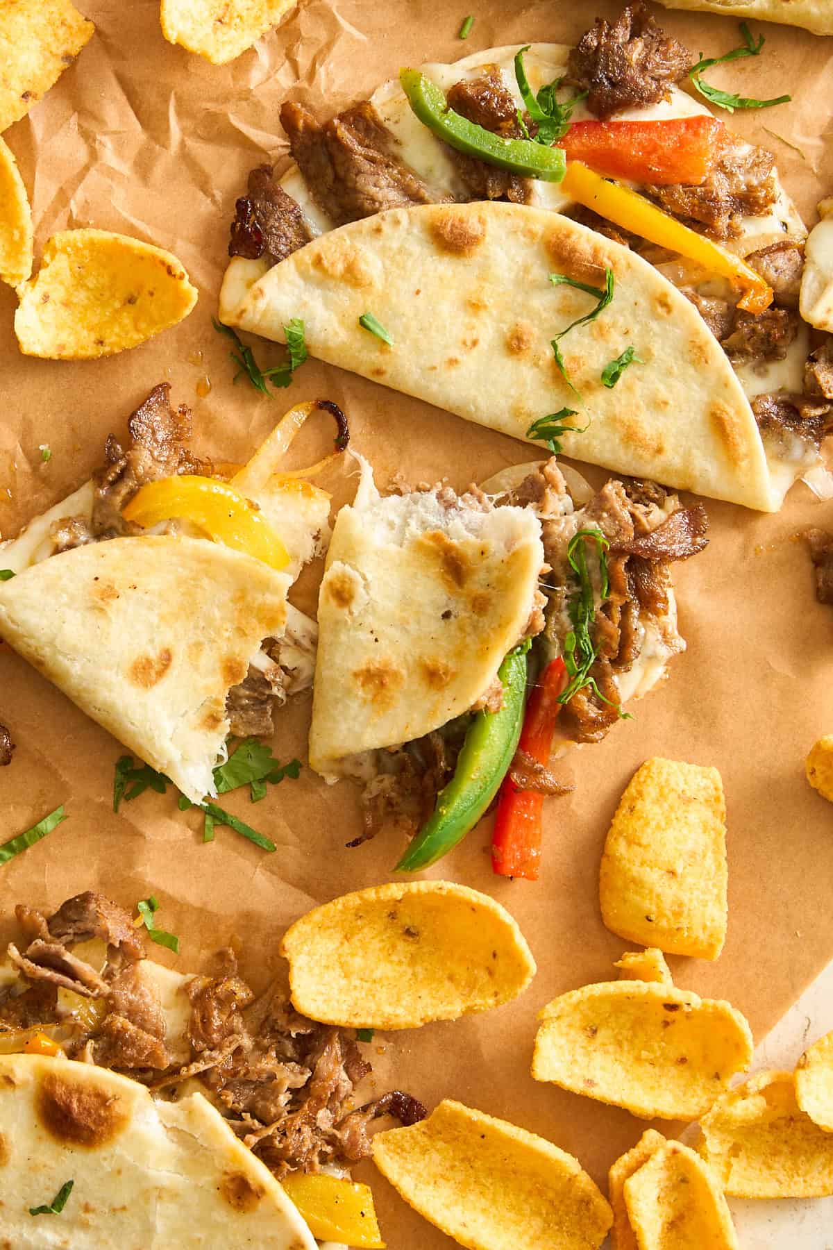 Soft tacos filled with shredded meat, yellow and green bell peppers, and herbs are arranged on brown parchment paper, surrounded by crunchy corn chips.