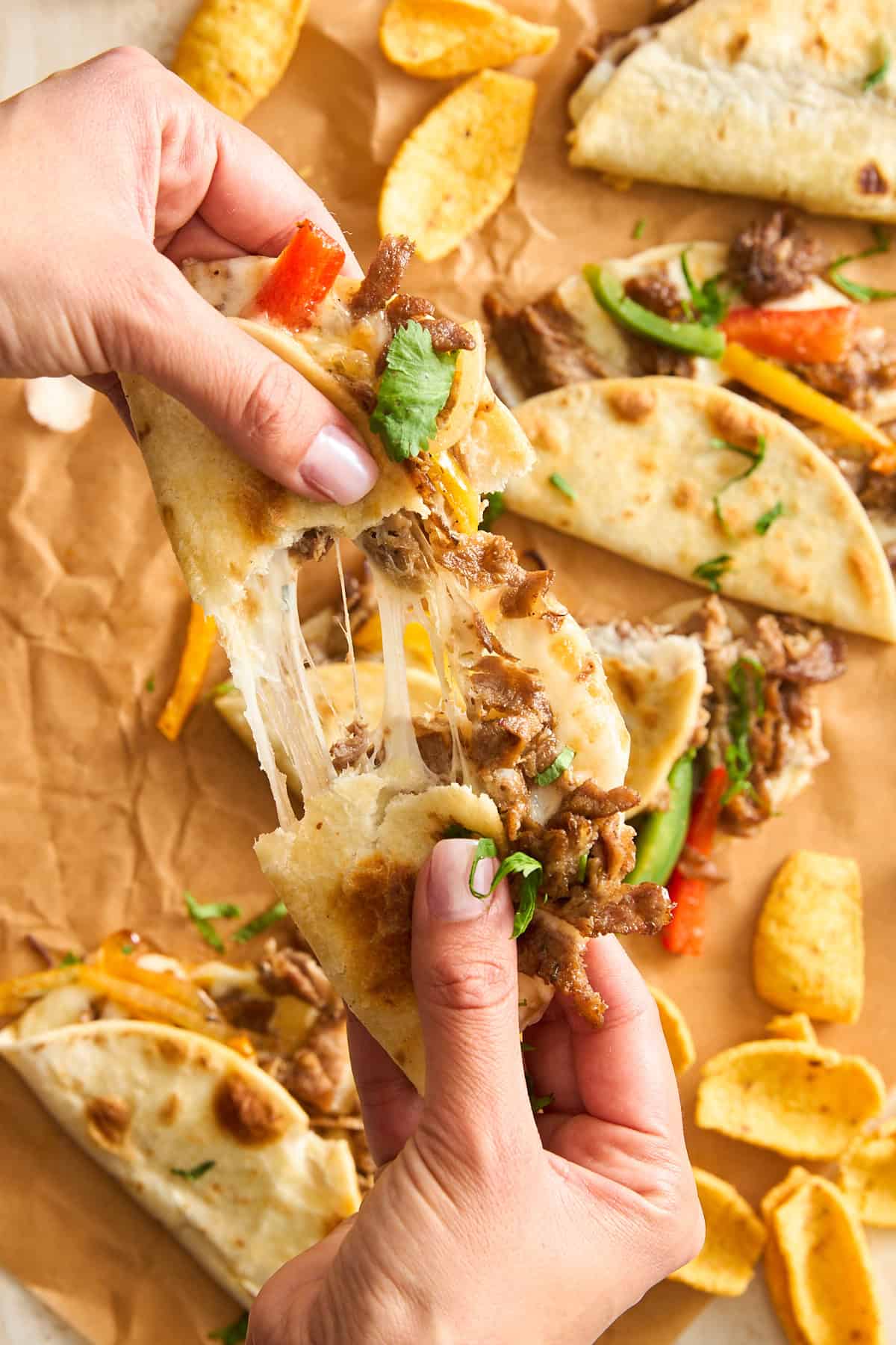 A person pulls apart a cheesy beef Philly cheesesteak taco with peppers and cilantro. Other quesadilla slices and curved corn chips are on brown parchment paper in the background.