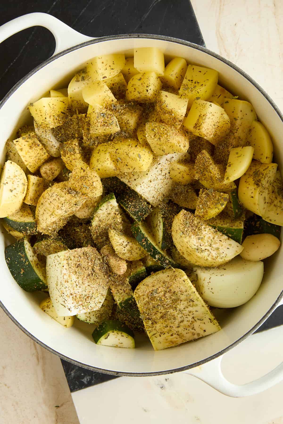 A white pot filled with chopped potatoes, zucchini, onions, and garlic cloves, all seasoned with a mix of dried herbs and black pepper, sits on a marble countertop.