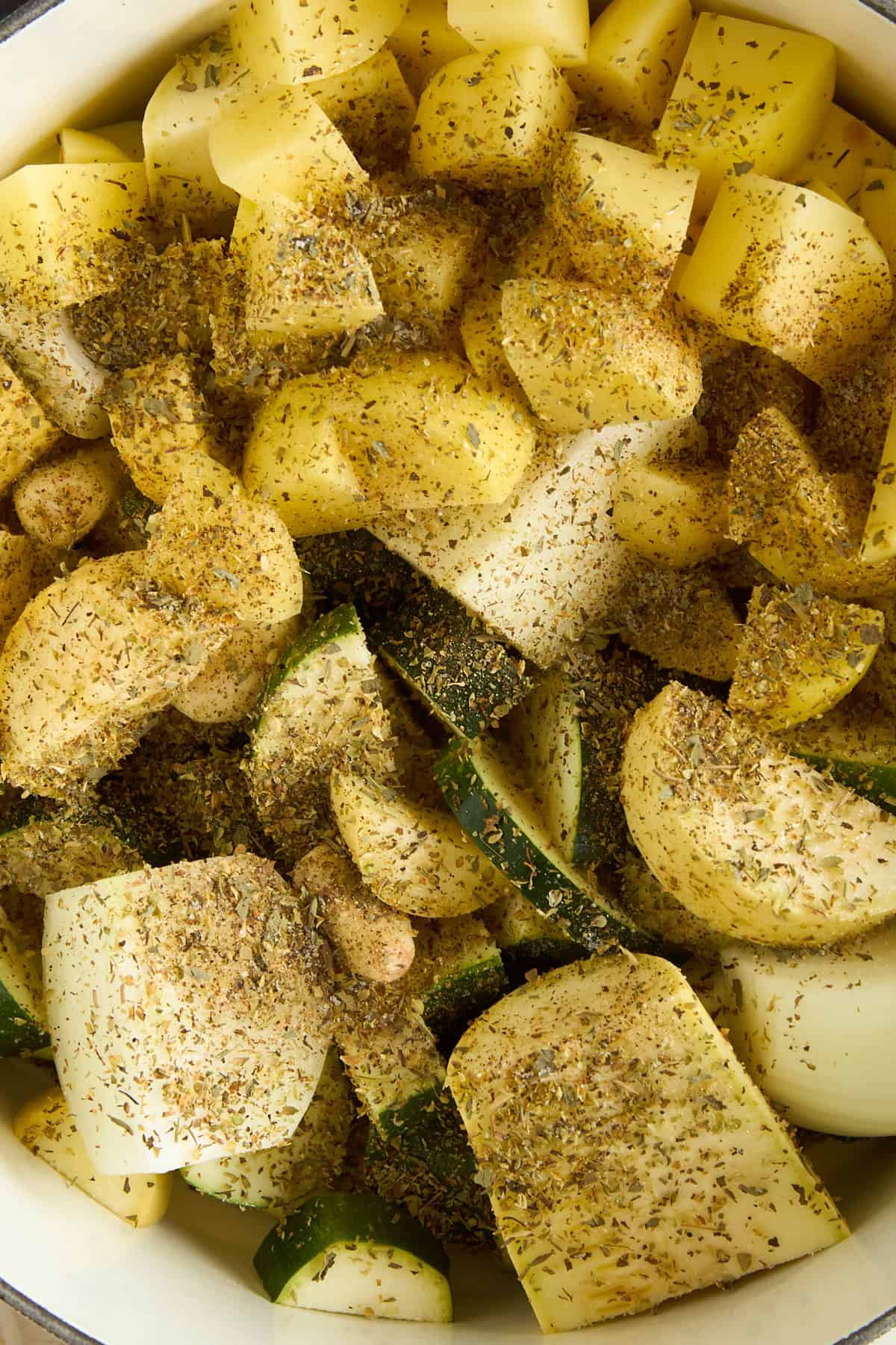 Chunks of potatoes, zucchini, and onions in a pot, generously sprinkled with dried herbs and spices, ready to be cooked.