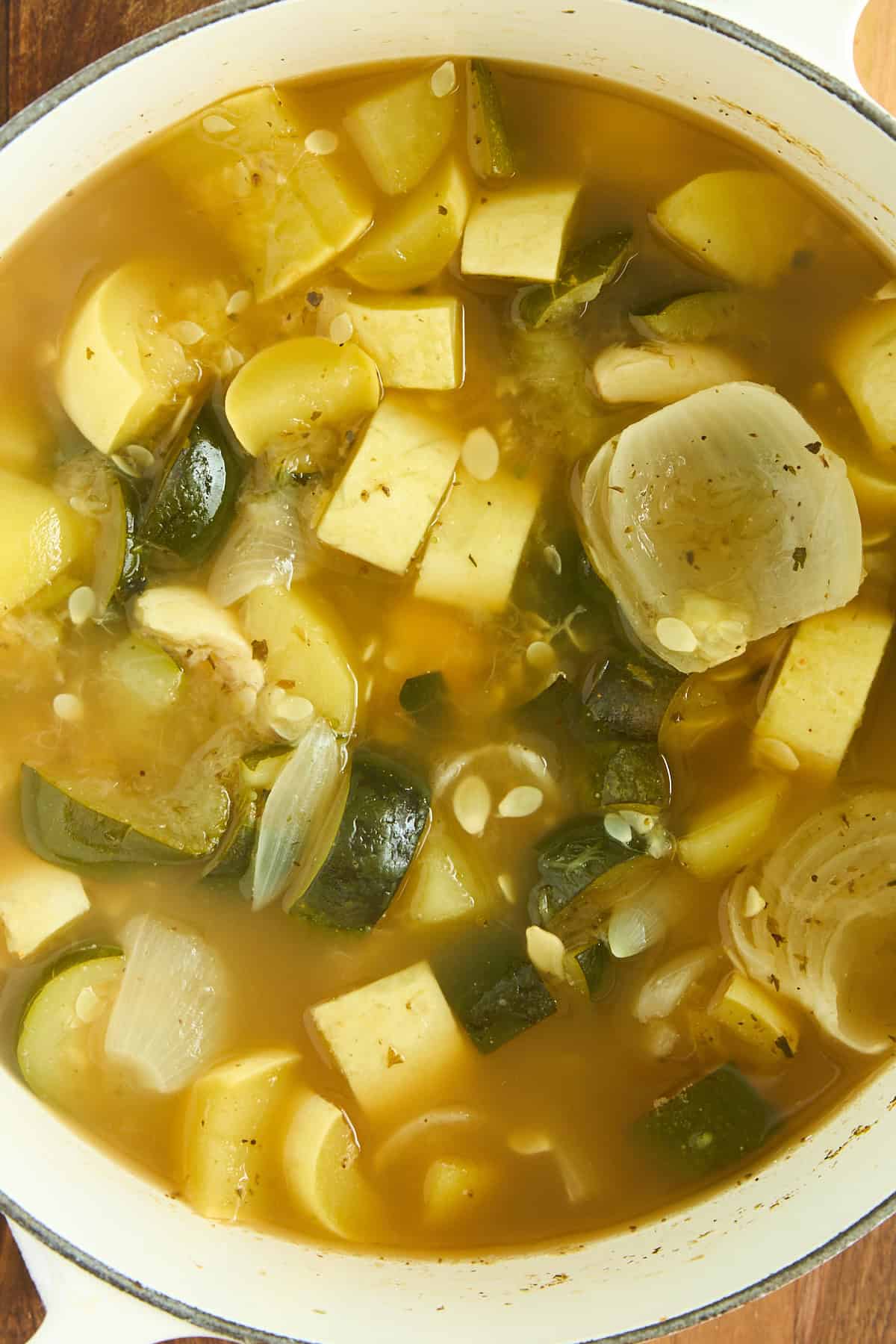 A close-up of a pot filled with vegetable soup, featuring chunks of onion, zucchini, and other vegetables in a clear broth.