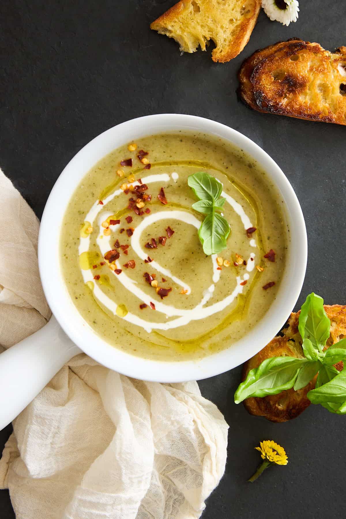 A bowl of creamy green soup garnished with a swirl of cream, chili flakes, olive oil, and fresh basil, served with toasted bread slices on a dark surface beside a light cloth and a yellow flower.