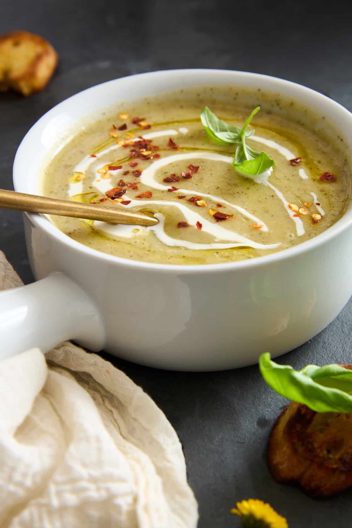 A creamy green soup in a white bowl, garnished with a swirl of cream, red pepper flakes, and fresh basil, with a gold spoon resting inside. A light cloth and toasted bread are nearby on a dark surface.