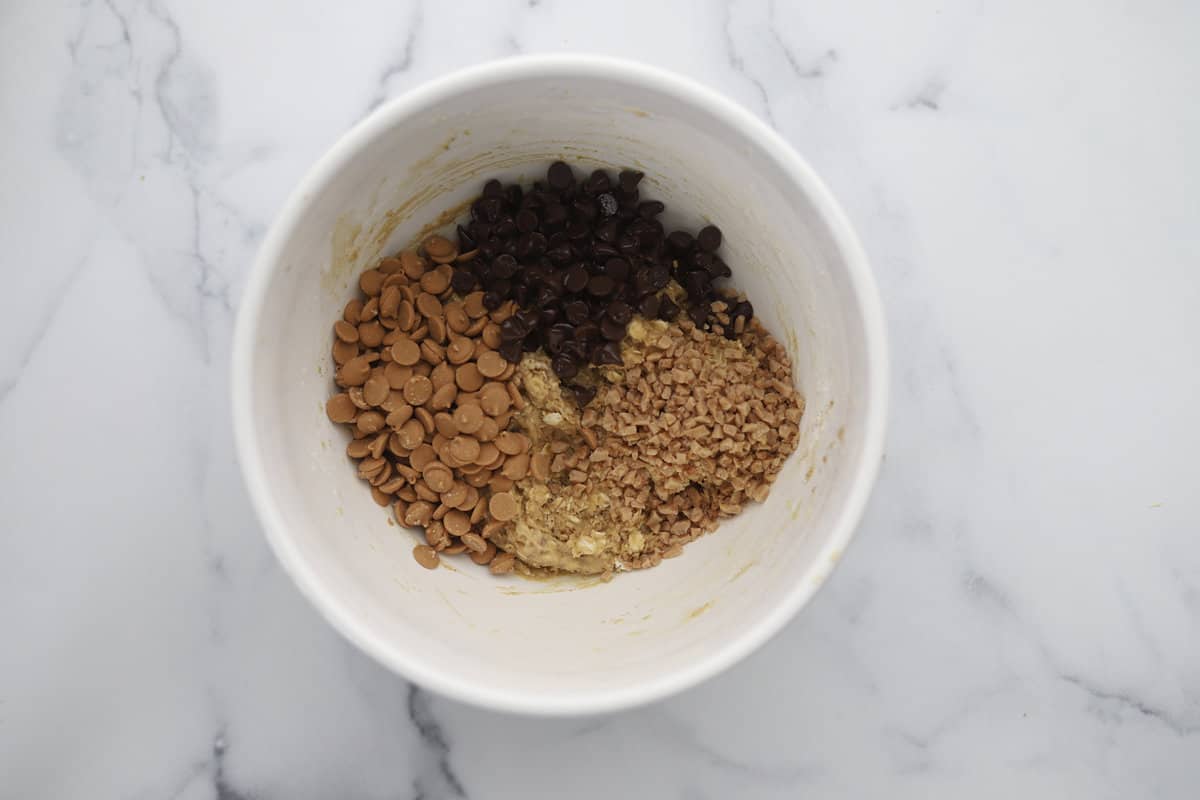 Chocolate chips, peanut butter chips, and toffee pieces being folded into cookie dough batter.