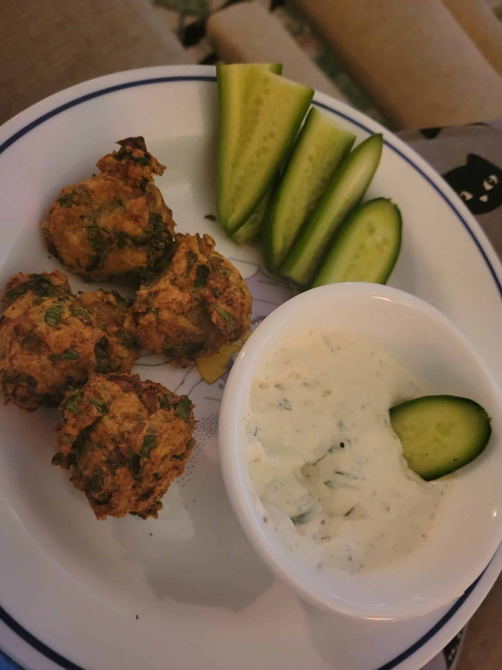 A plate with three falafel balls, four cucumber slices, and a small bowl of white yogurt sauce garnished with herbs and a cucumber slice. The food is arranged neatly on a white plate with a blue rim.