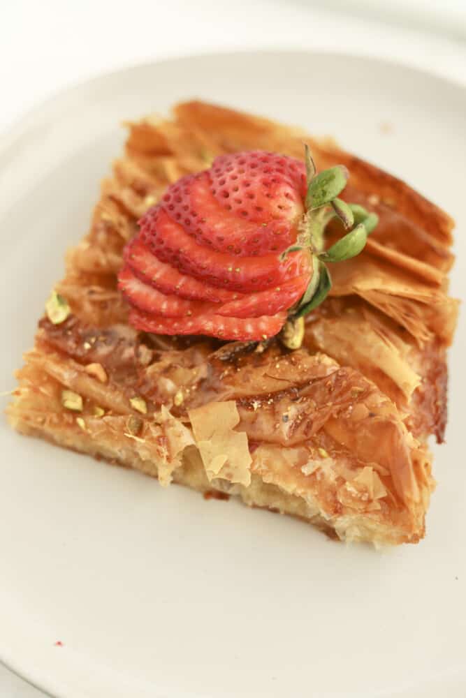 close up image of a white plate holding a piece of a baked crinkle phyllo dough recipe topped with a few pistachios and a sliced strawberry