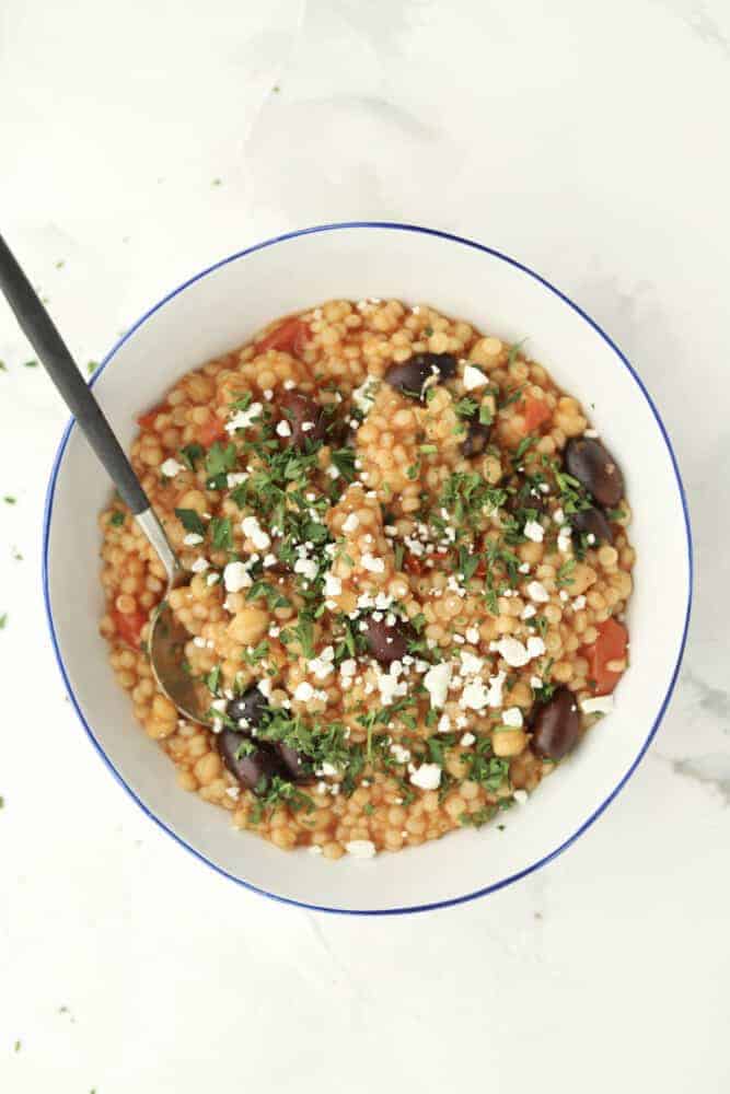 a bowl of Greek couscous topped with crumbled feta and fresh herbs with a spoon sticking out 