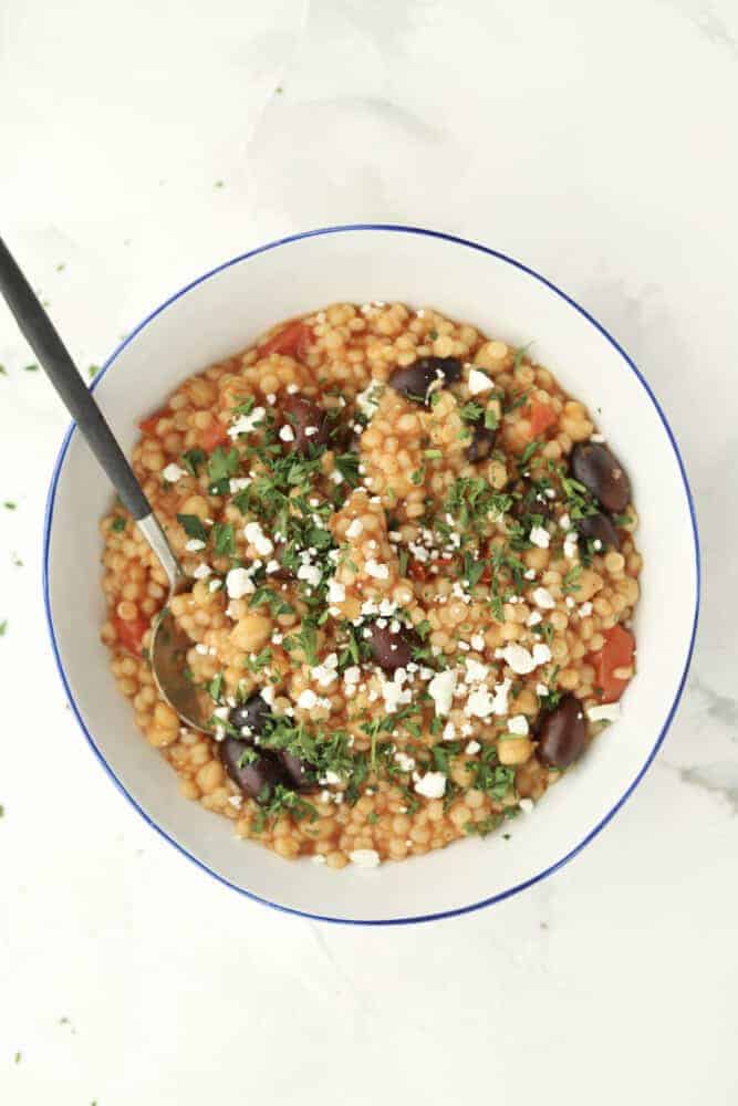 a bowl of Greek couscous topped with crumbled feta and fresh herbs with a spoon sticking out