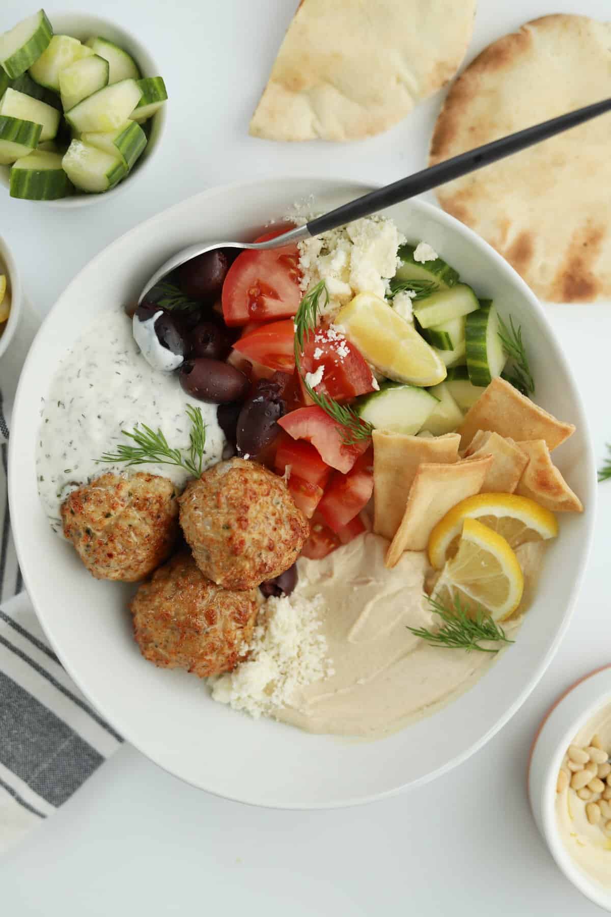 chicken kofta in a bowl with hummus, tomatoes, cucumbers, and tzaziki sauce.